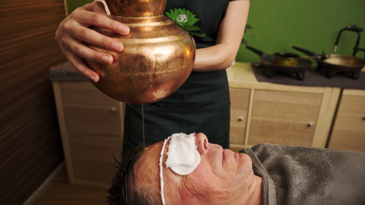 Therapist pouring warm oil from a copper vessel during a Shirodhara treatment for a male client